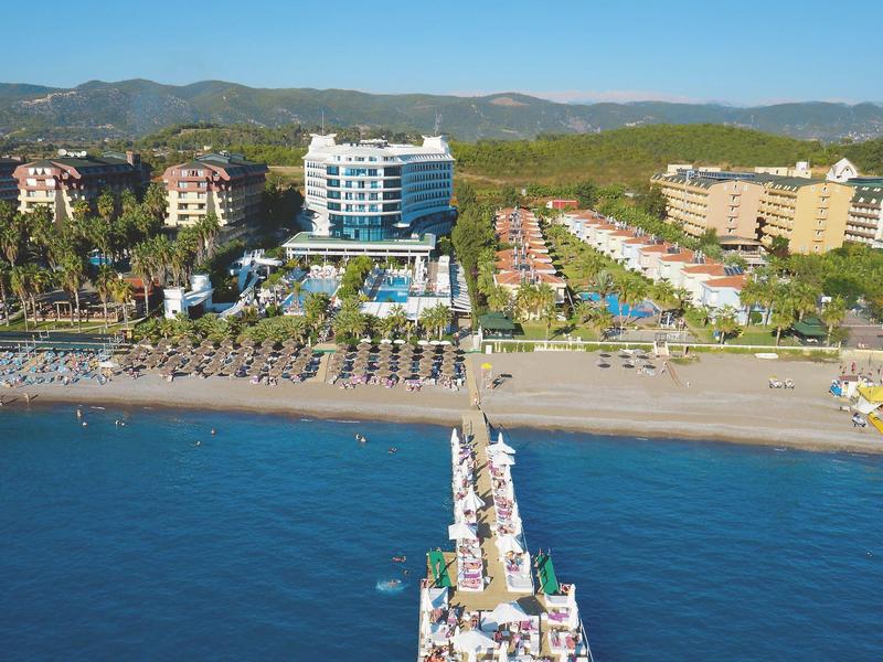 Pohled na pláž s dlouhým molem a několika hotelovými budovami v pozadí pod jasnou oblohou.