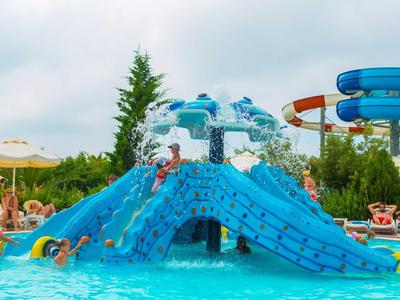 Buntes Wasserrutschen-Spielgerät mit Kletterwand in einem Außenpool, umgeben von Sonnenschirmen und Bäumen.
