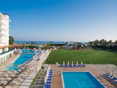 Blick auf Hotelpool mit Liegestühlen, Rasenfläche und Meer im Hintergrund unter blauem Himmel.