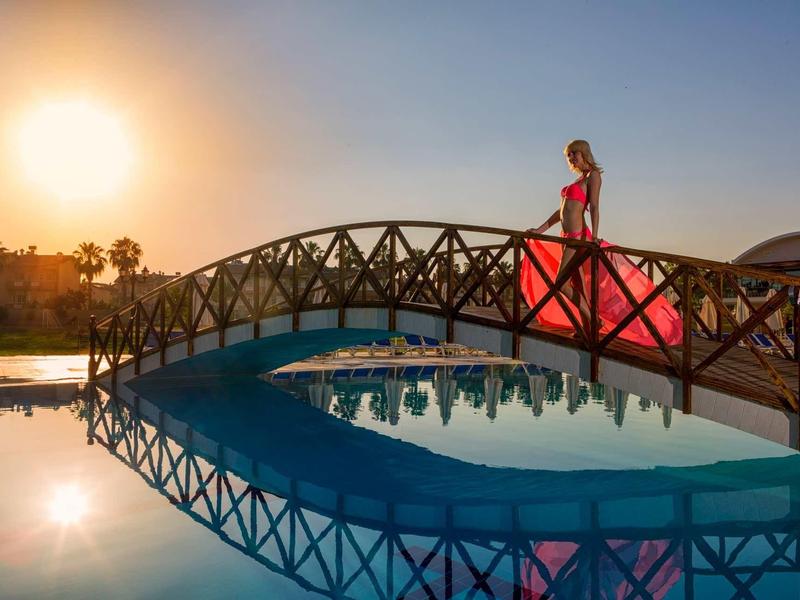 Une femme en robe rouge traverse un pont au-dessus d'une piscine au coucher du soleil.