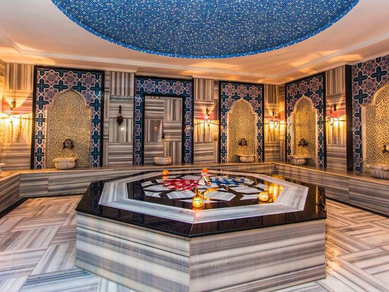 Hammam luxueux avec mosaïques, marbre et bassin central décoré.