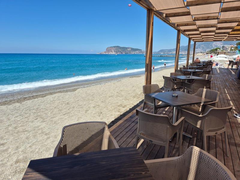 Restaurante en la playa con mesas y sillas de madera, vista al mar azul y cielo despejado.