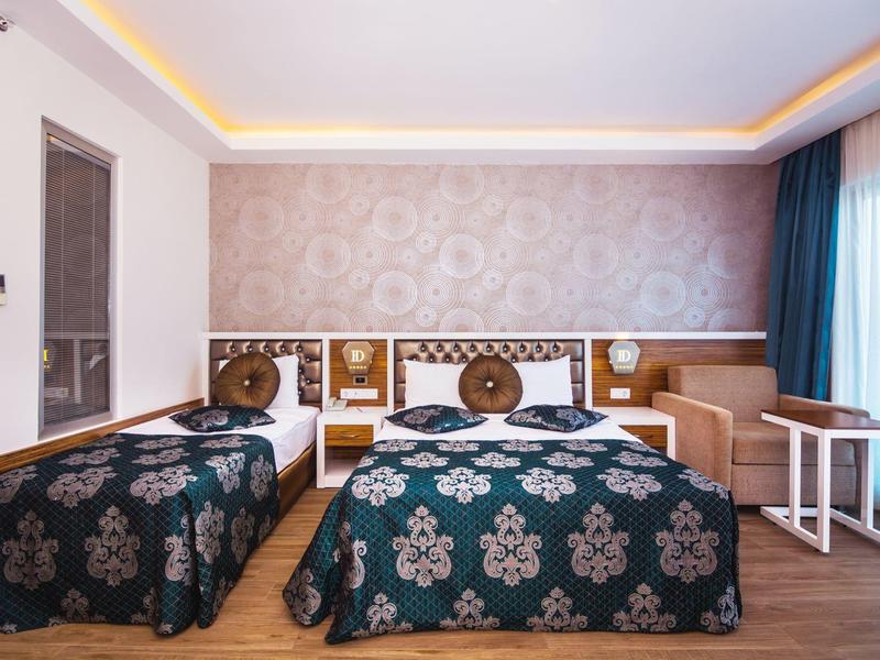 Habitación de hotel luminosa con dos camas, mantas estampadas, sofá y grandes ventanas con cortinas.