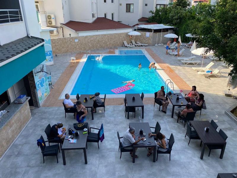 Hotel pool with guests seated at tables and sun loungers outdoors.