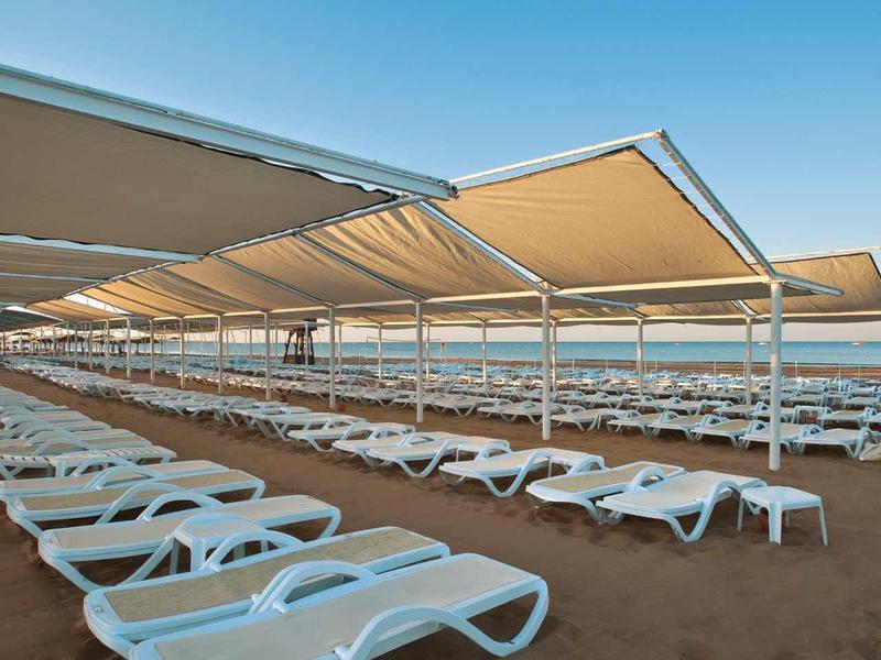 Multiple white lounge chairs arranged in rows on the beach under large sunshades.