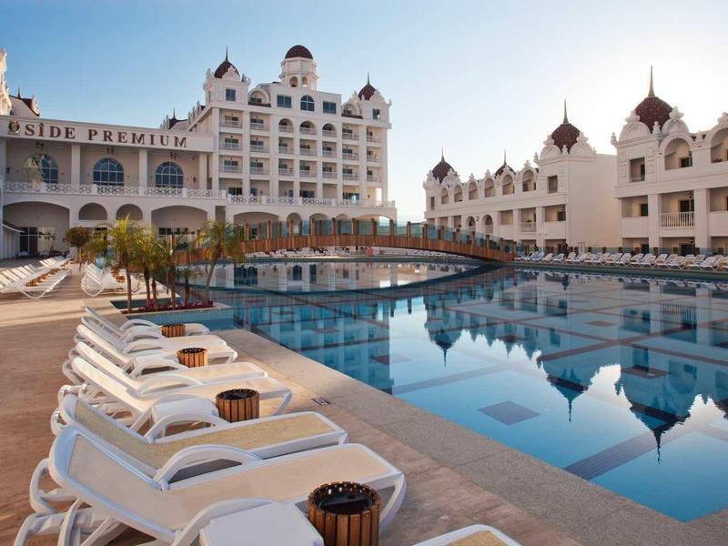 Large outdoor pool with sun loungers in front of a white hotel with turrets under clear sky.