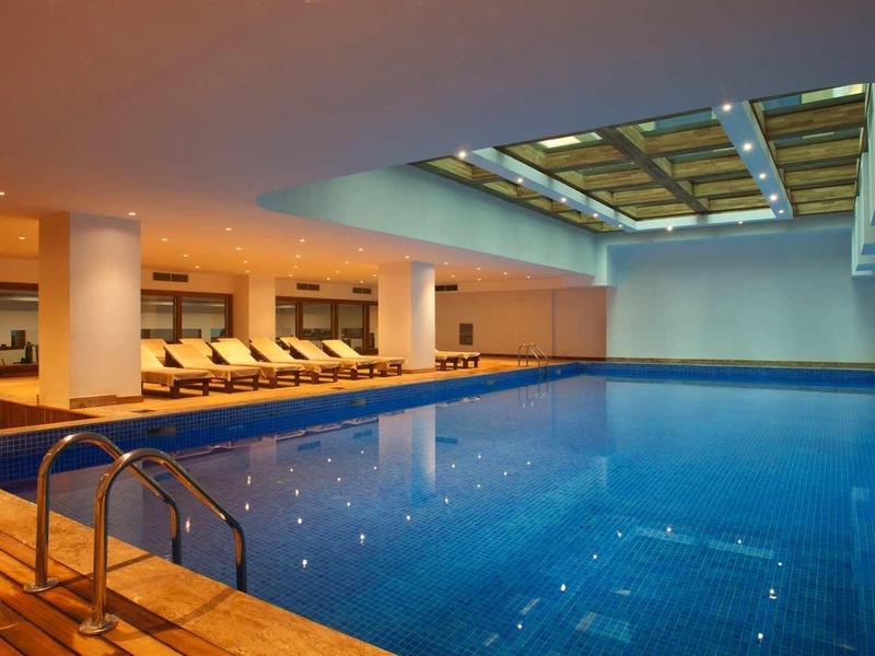 Modern indoor pool with lounge chairs and large glass roof in a hotel.