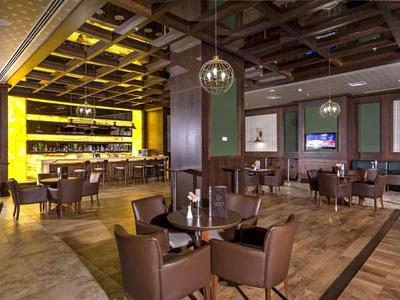 Modern restaurant with brown leather chairs, wooden tables, and a yellow-lit bar area.