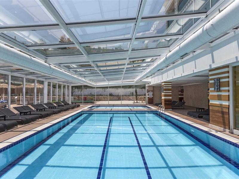 Indoor pool with glass roof and lounge chairs beside the water in a modern hotel.