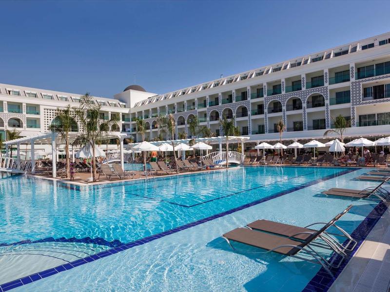 Large hotel with swimming pool, lounge chairs, and umbrellas under clear sky