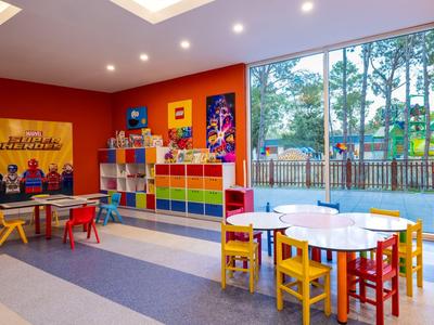 Bunter Kinderspielraum mit Tischen, Stühlen und großen Fenstern, durch die Bäume sichtbar sind.
