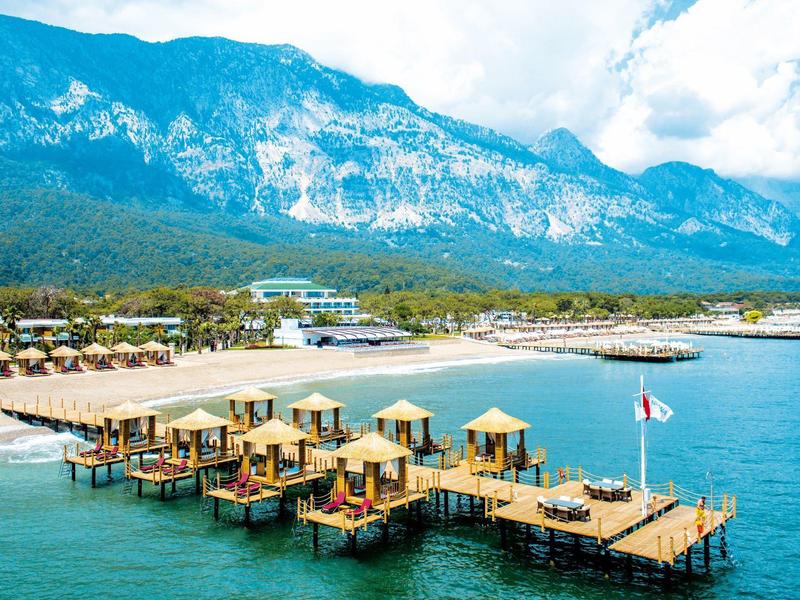 Wooden platforms with sun umbrellas extend into the blue sea against a mountain with cloudy sky.