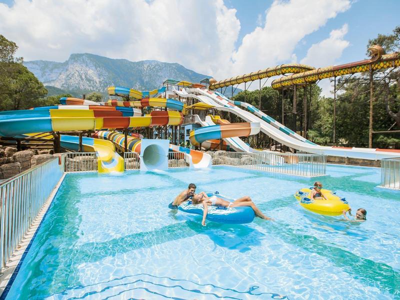 Water park with colorful water slides and people on inflatables in a pool with mountains in the background.