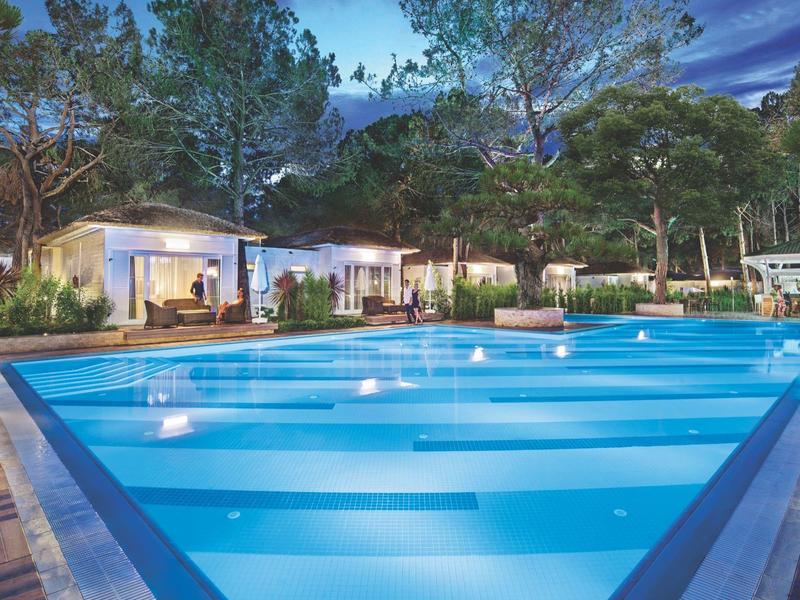 Illuminated outdoor pool in front of a modern hotel building at dusk, surrounded by trees.