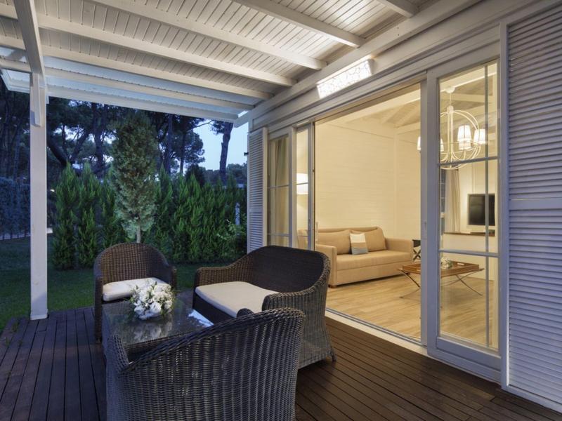 Covered terrace with wicker seating area, view into the bright living room of a hotel.