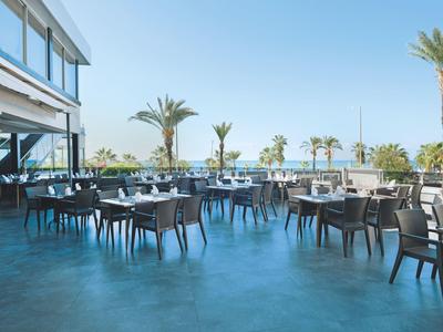 Leere Außenterrasse mit Tischen und Stühlen neben einem Hotel mit Blick auf Palmen und blauen Himmel.