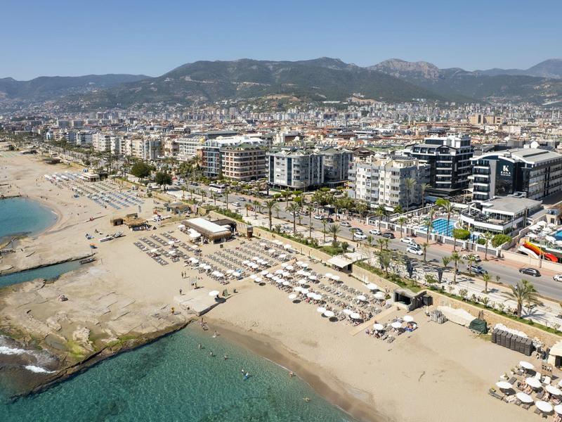 Blick auf einen weitläufigen Strand mit Liegestühlen und dahinter einer Stadt mit Bergen im Hintergrund.