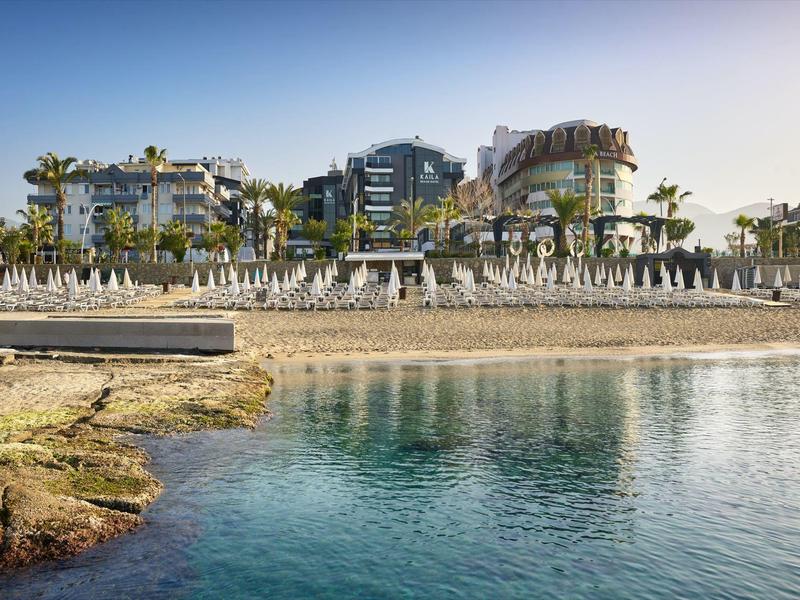Strand mit Sand, Liegestühlen und Handtüchern vor Hotels und Palmen bei klarem Himmel.