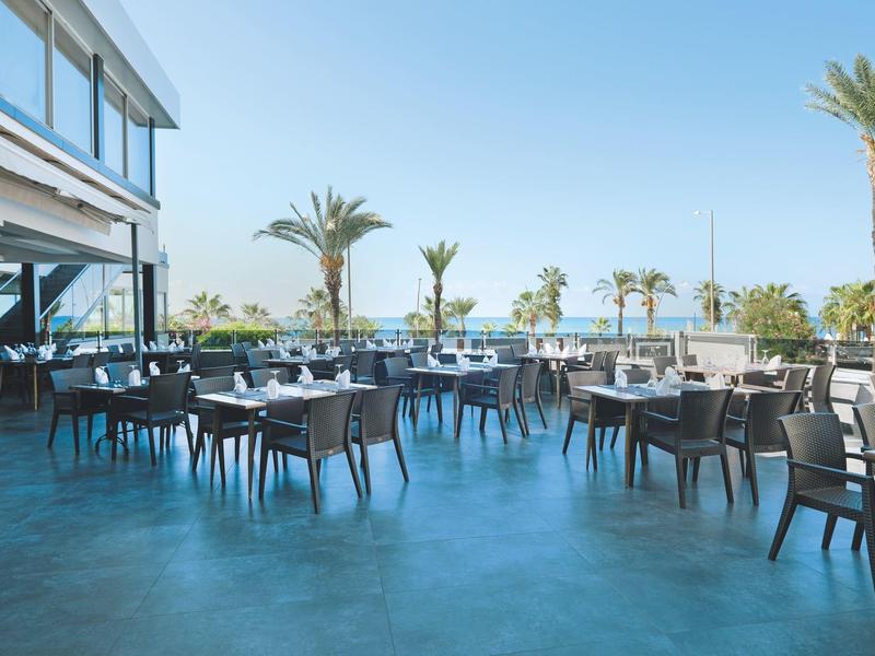 Leere Außenterrasse mit Tischen und Stühlen neben einem Hotel mit Blick auf Palmen und blauen Himmel.
