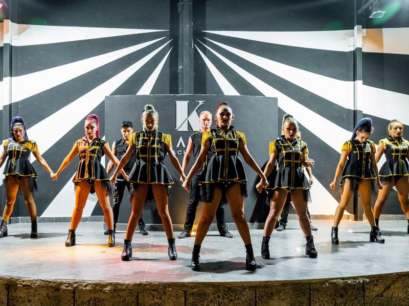 Gruppe von Tänzerinnen in schwarzen und goldenen Kostümen auf einer Bühne mit schwarz-weißer geometrischer Wand.