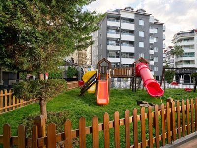 Playground with swing and slides in front of residential buildings and wooden fence in green area.