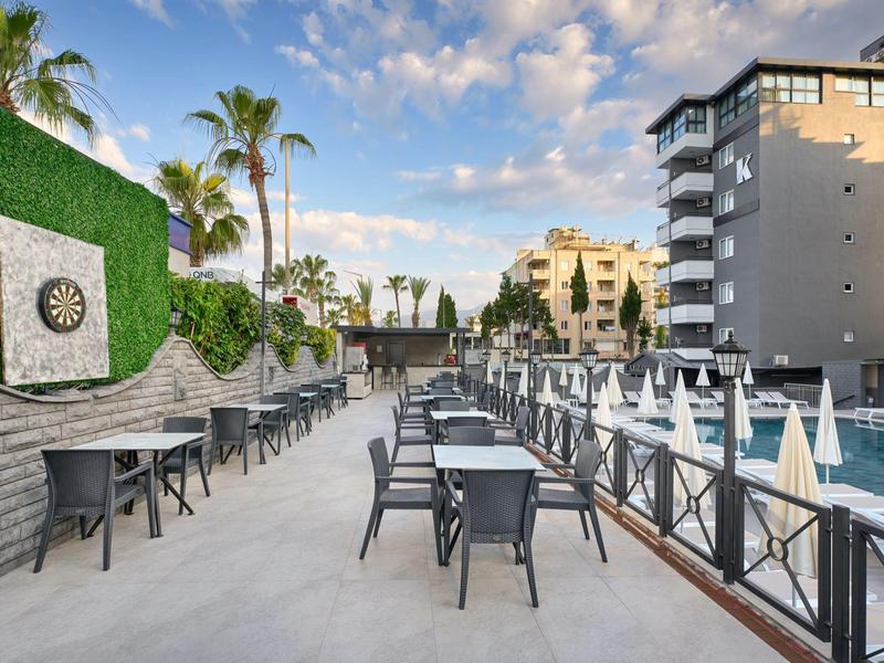 Modern outdoor area with pool, tables, and chairs next to a hotel building under blue sky.