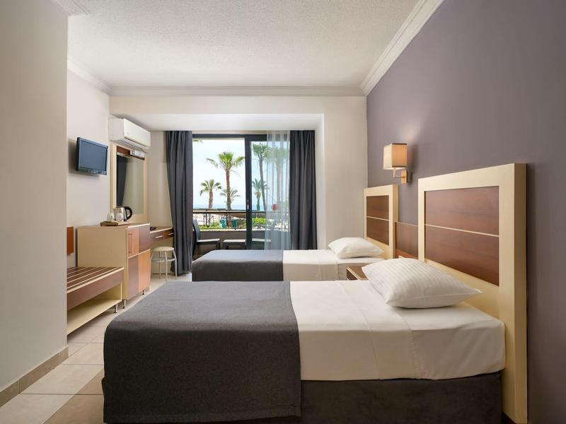 Modern hotel room with two single beds, TV, and balcony overlooking the sea.