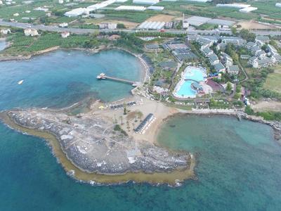 Coastal resort with pools, green areas, pier, and surrounding blue sea, viewed from above.