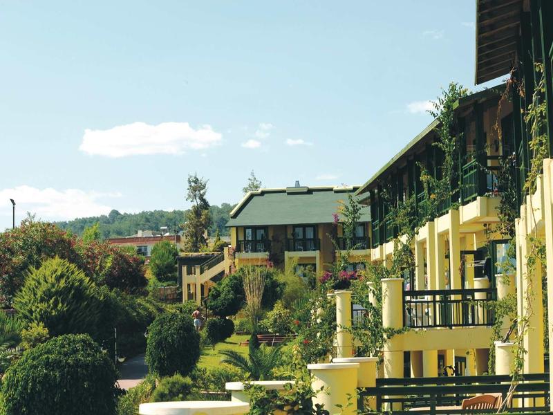 Yellow buildings with balconies surrounded by green trees and bushes under a partly cloudy sky.