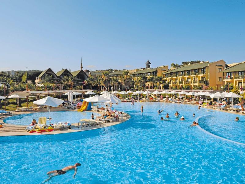 Large outdoor pool with people swimming and relaxing near resort buildings under a clear sky.