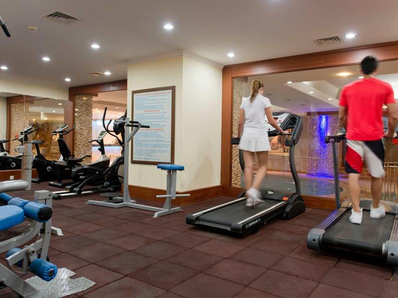 Two people running on treadmills in a hotel gym with white walls and brown carpet flooring.