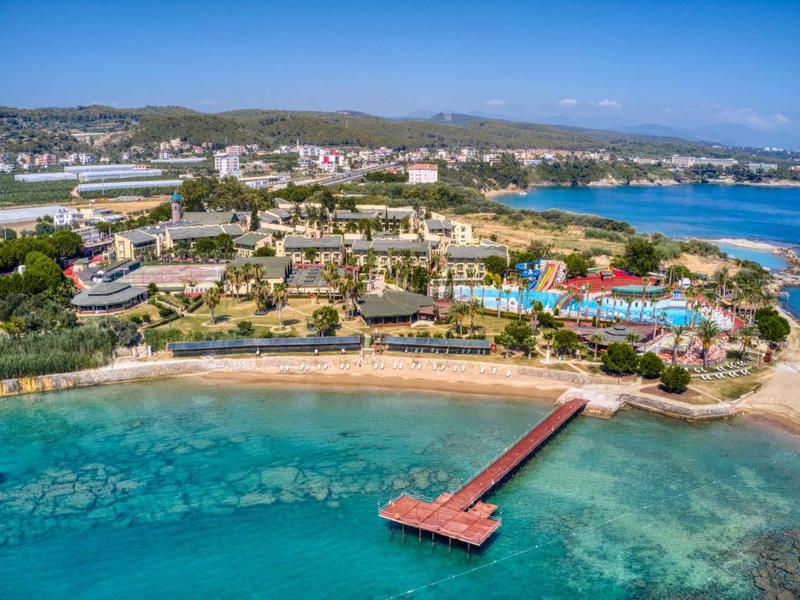 Coastal resort with beach, pier, pools, and buildings by clear blue sea and hills in the background.