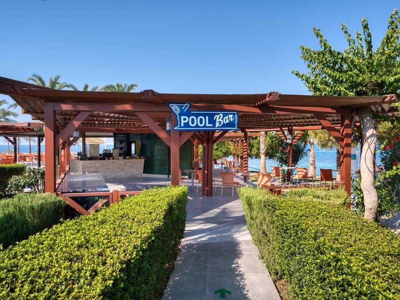 Wooden pool bar with shaded seating, surrounded by green hedges and tropical plants.