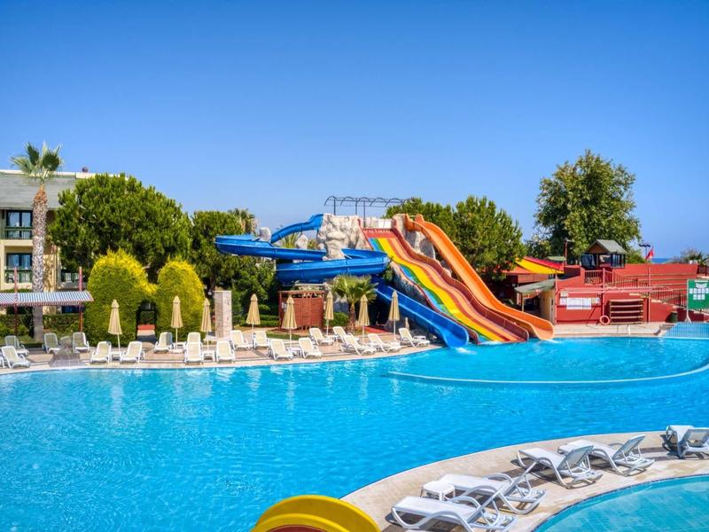 Bright outdoor pool with colorful slides and sun loungers near hotel buildings under clear blue sky.