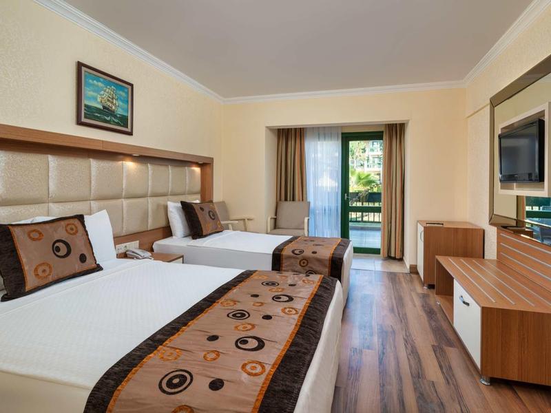 Bright hotel room with two beds, wooden floor, TV, and balcony doors letting in natural light.