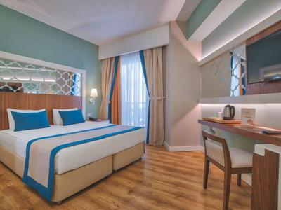 Camera d'hotel luminosa con letto matrimoniale, scrivania, sedia e grandi tende alla finestra.