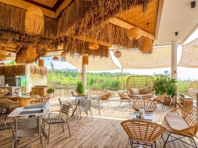 Offenes Café mit Holzboden, geflochtenen Stühlen, Holzdecke und Blick auf grüne Landschaft.