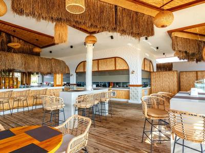 Bar luminosa e aperta con sgabelli alti, mobili in legno e rattan, soffitto decorativo e atmosfera accogliente.