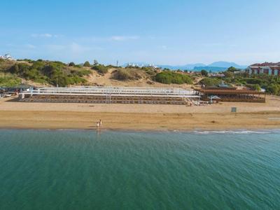 Spiaggia con sabbia e mare calmo, edifici bassi e vegetazione sullo sfondo sotto un cielo azzurro.