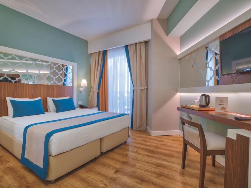 Camera d'hotel luminosa con letto matrimoniale, scrivania, sedia e grandi tende alla finestra.