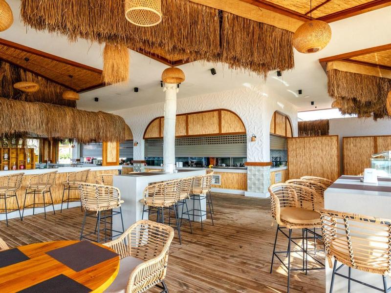 Bar luminosa e aperta con sgabelli alti, mobili in legno e rattan, soffitto decorativo e atmosfera accogliente.