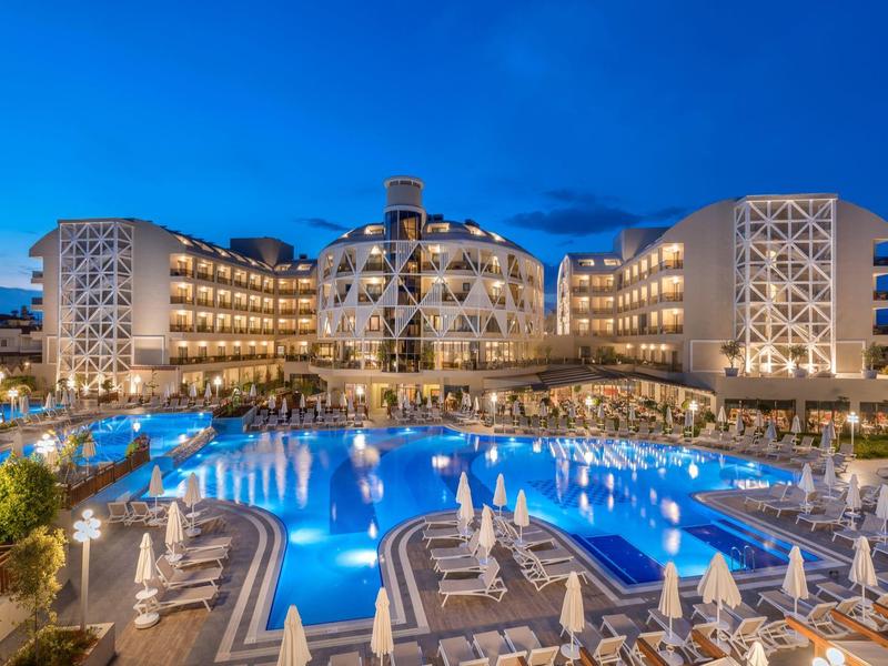 Complesso alberghiero moderno di notte con piscina illuminata e lettini in primo piano.