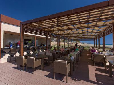 Zastřešená terasa restaurace se židlemi a stoly, v pozadí výhled na moře.