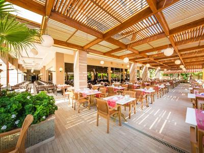 Helles, luftiges Restaurant mit Holzboden, gedeckten Tischen und vielen Pflanzen unter Pergola.