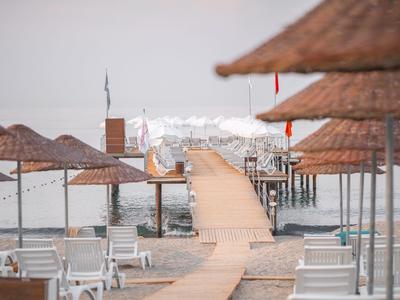 Strand mit Holzsteg, Liegestühlen und Schirmen, dahinter ruhiges Wasser und verschwommene Boote.