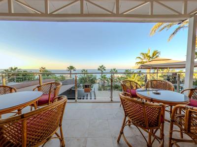 Terras met tafels en stoelen met uitzicht op palmbomen en zee bij zonsondergang.