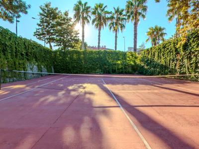 Leeg tennisveld met rode bodem omgeven door palmbomen en dichte groene klimop onder een blauwe lucht.