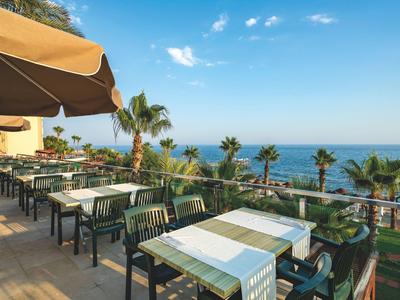 Terras met tafels en stoelen met uitzicht op palmbomen en de blauwe zee bij een heldere lucht.