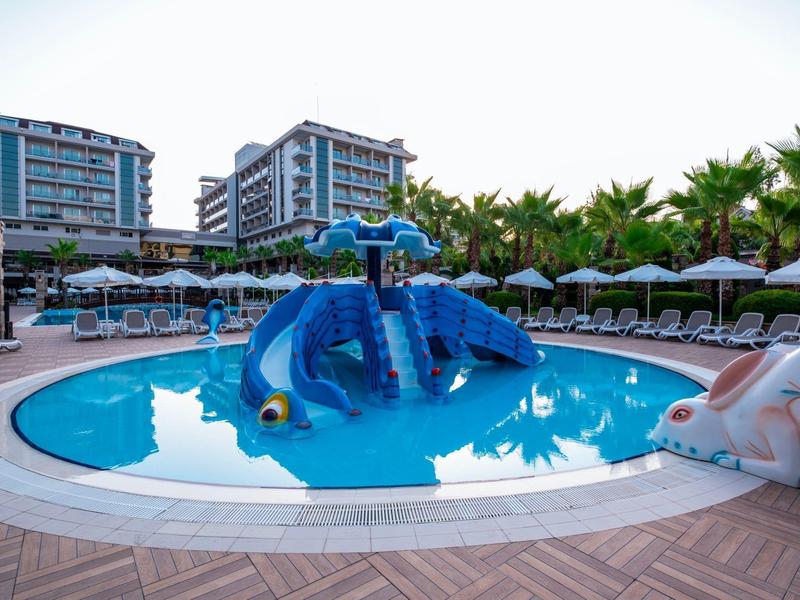 Hotel pool with blue dolphin slide, wooden deck, and umbrellas near tall buildings and palm trees.
