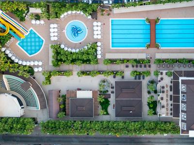 Luftaufnahme eines Hotelpools mit Wasserrutschen, Schwimmbecken, Sonnenschirmen und umgebenden Grünflächen.